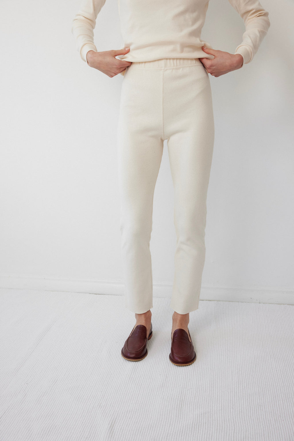 A person wears Wol Hide’s Easy Legging in Natural, paired with a matching top. Only their body from shoulders down is visible. They stand on a light carpet in brown loafers against a white wall.
