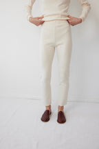A person wears Wol Hide’s Easy Legging in Natural, paired with a matching top. Only their body from shoulders down is visible. They stand on a light carpet in brown loafers against a white wall.