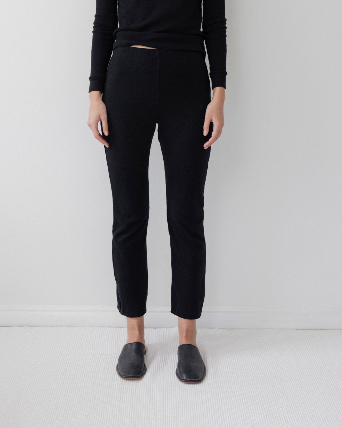 A person stands against a plain white wall, wearing Wol Hide's Easy Legging in Black, paired with a black long-sleeve top and dark slip-on shoes. Their arms rest at their sides on a light-colored floor.