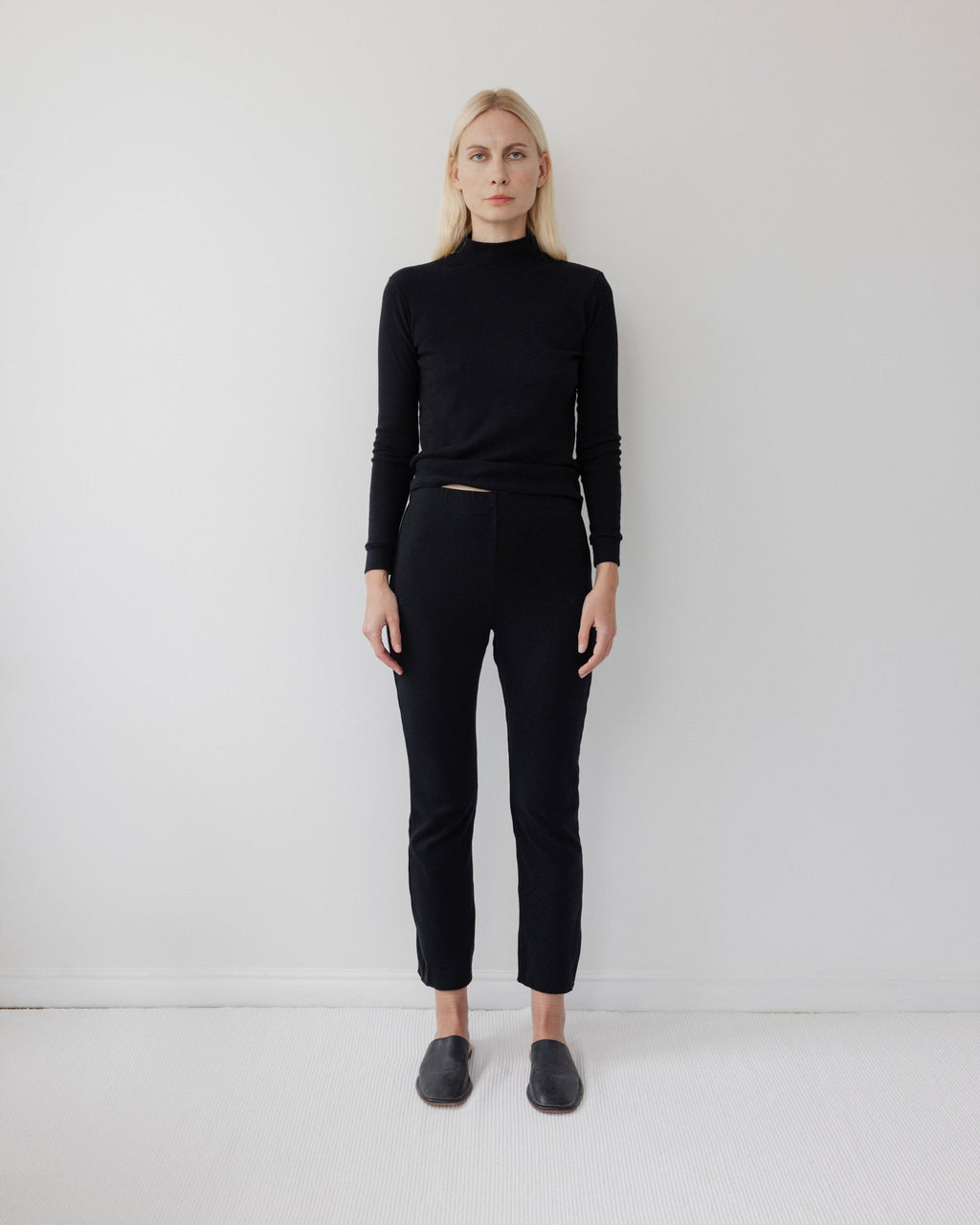 A person with long blonde hair stands against a plain white wall, wearing Wol Hide's Easy Legging in Black with a fitted black turtleneck and black slip-on shoes, arms at their sides and a neutral expression.