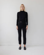 A person with long blonde hair stands against a plain white wall, wearing Wol Hide's Easy Legging in Black with a fitted black turtleneck and black slip-on shoes, arms at their sides and a neutral expression.