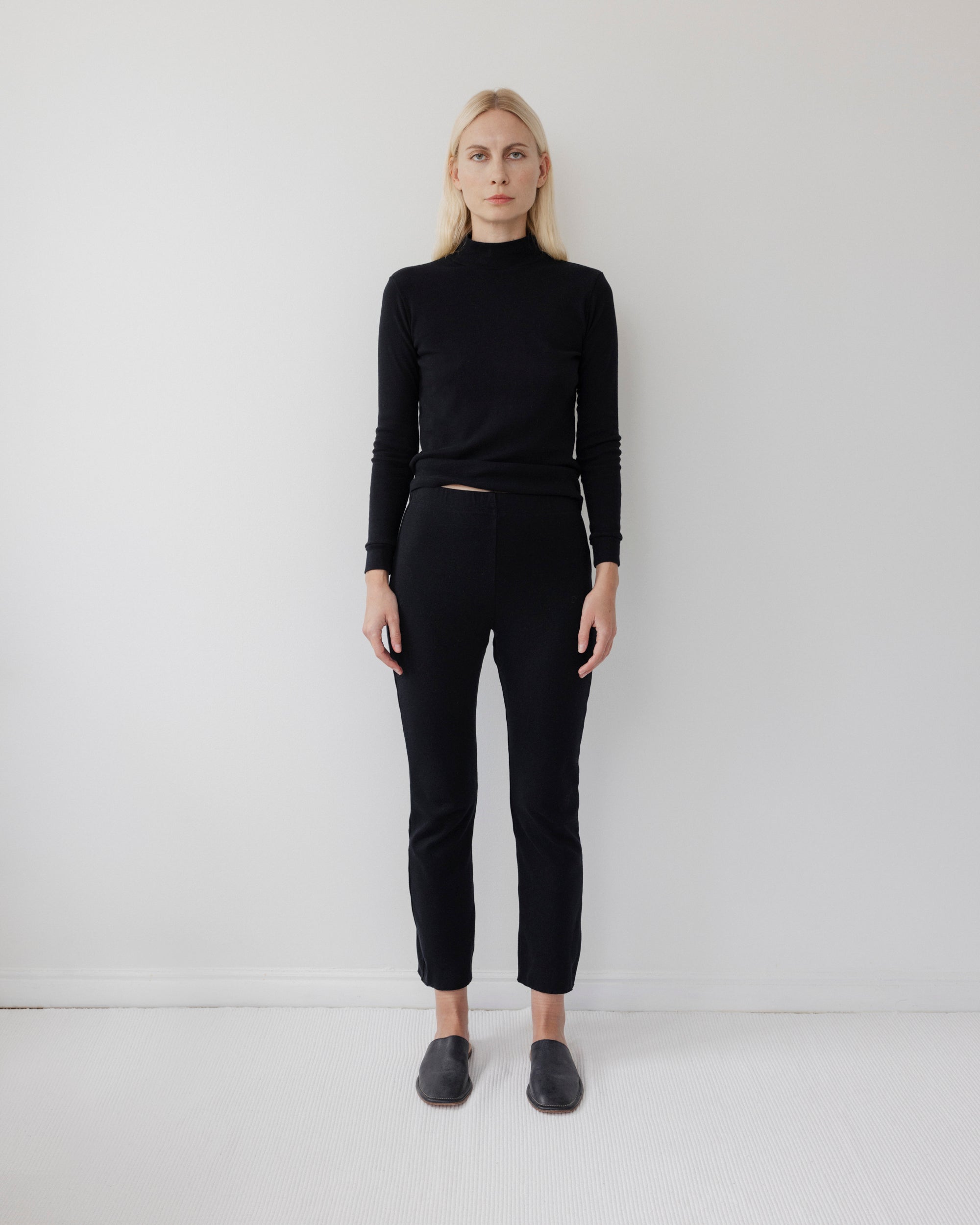 A person with long blonde hair stands against a plain white wall, wearing Wol Hide's Easy Legging in Black with a fitted black turtleneck and black slip-on shoes, arms at their sides and a neutral expression.