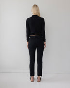 A person with long blonde hair stands barefoot, back to the camera, wearing Wol Hide's Easy Legging in Black and a black long-sleeve top, against a plain light-colored wall.