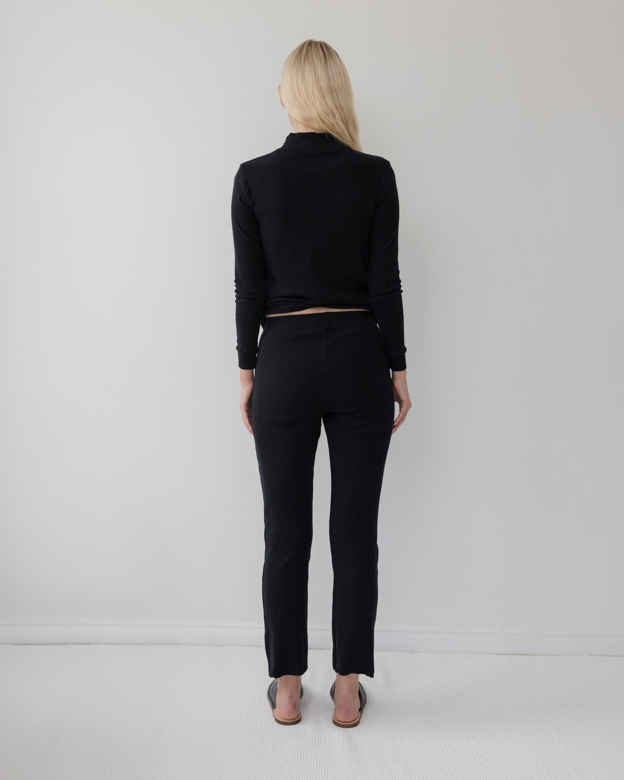 A person with long blonde hair stands barefoot, back to the camera, wearing Wol Hide's Easy Legging in Black and a black long-sleeve top, against a plain light-colored wall.