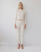 A person with long blonde hair stands barefoot against a plain white wall, wearing Wol Hide's Easy Legging in Natural and a light beige long-sleeve top, looking straight ahead with a neutral expression.
