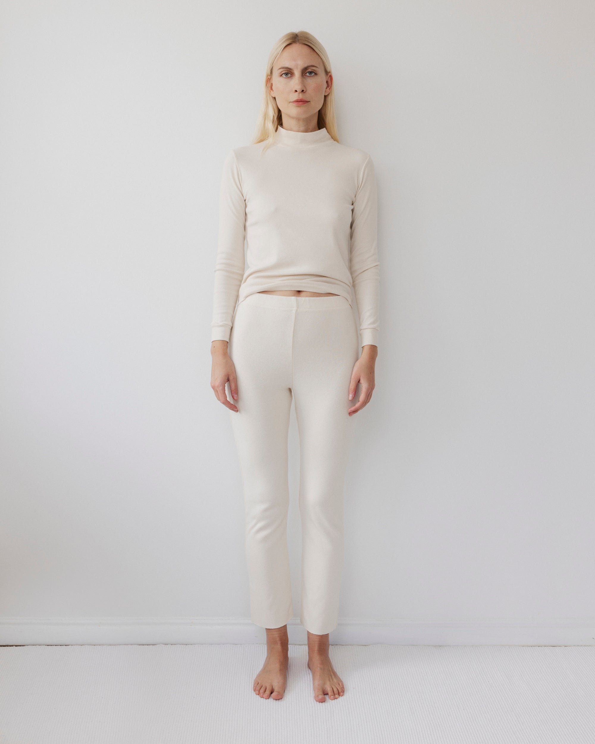 A person with long blonde hair stands barefoot against a plain white wall, wearing Wol Hide's Easy Legging in Natural and a light beige long-sleeve top, looking straight ahead with a neutral expression.