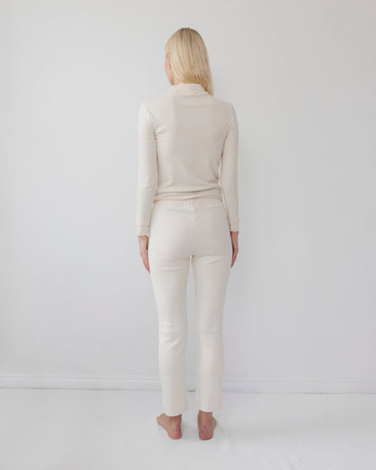 A woman with long blonde hair stands barefoot, facing away, wearing a light-colored, form-fitting ensemble featuring the Easy Legging in Natural from Wol Hide. The leggings are crafted from USA-made organic cotton and set against a plain white background.