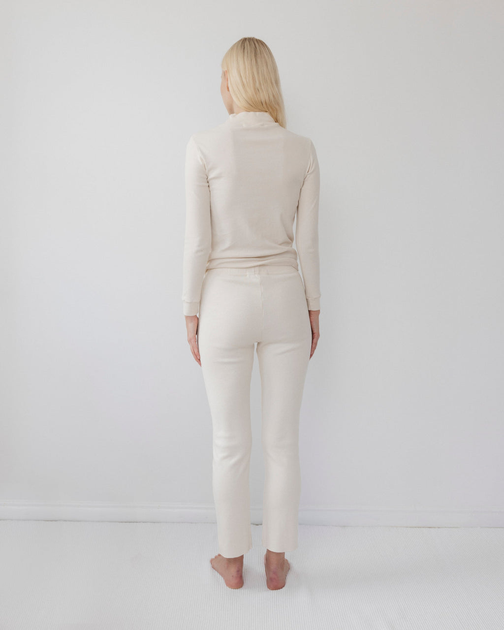 A person with long blonde hair stands barefoot facing a white wall, wearing the Wol Hide Easy Legging in Natural and a matching cream-colored fitted long-sleeve top.