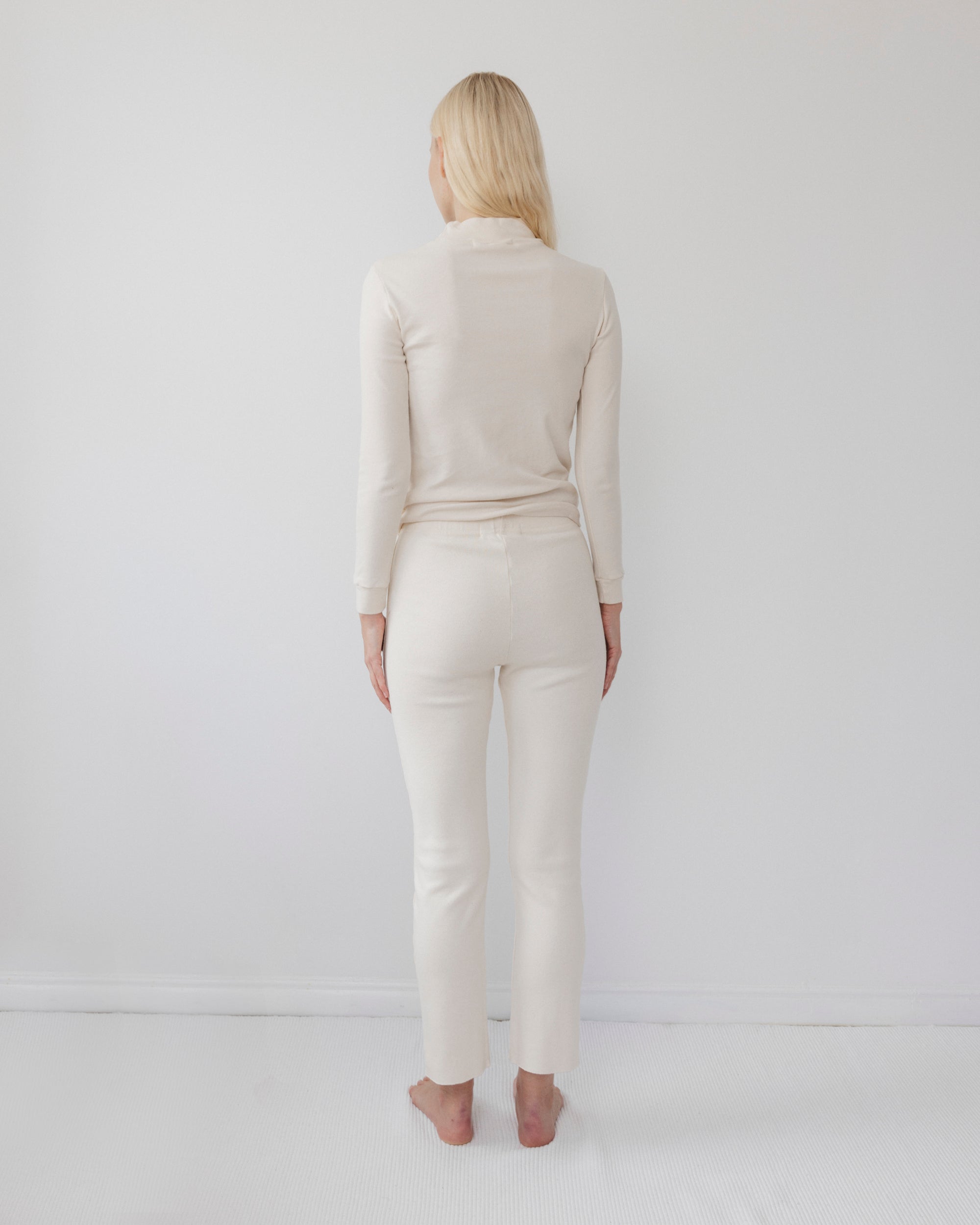 A person with long blonde hair stands barefoot facing a white wall, wearing the Wol Hide Easy Legging in Natural and a matching cream-colored fitted long-sleeve top.