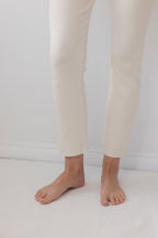 A person stands barefoot on a white textured surface, wearing Wol Hide's Easy Legging in Natural—organic cotton leggings that end above the ankles. A plain white wall forms the background.