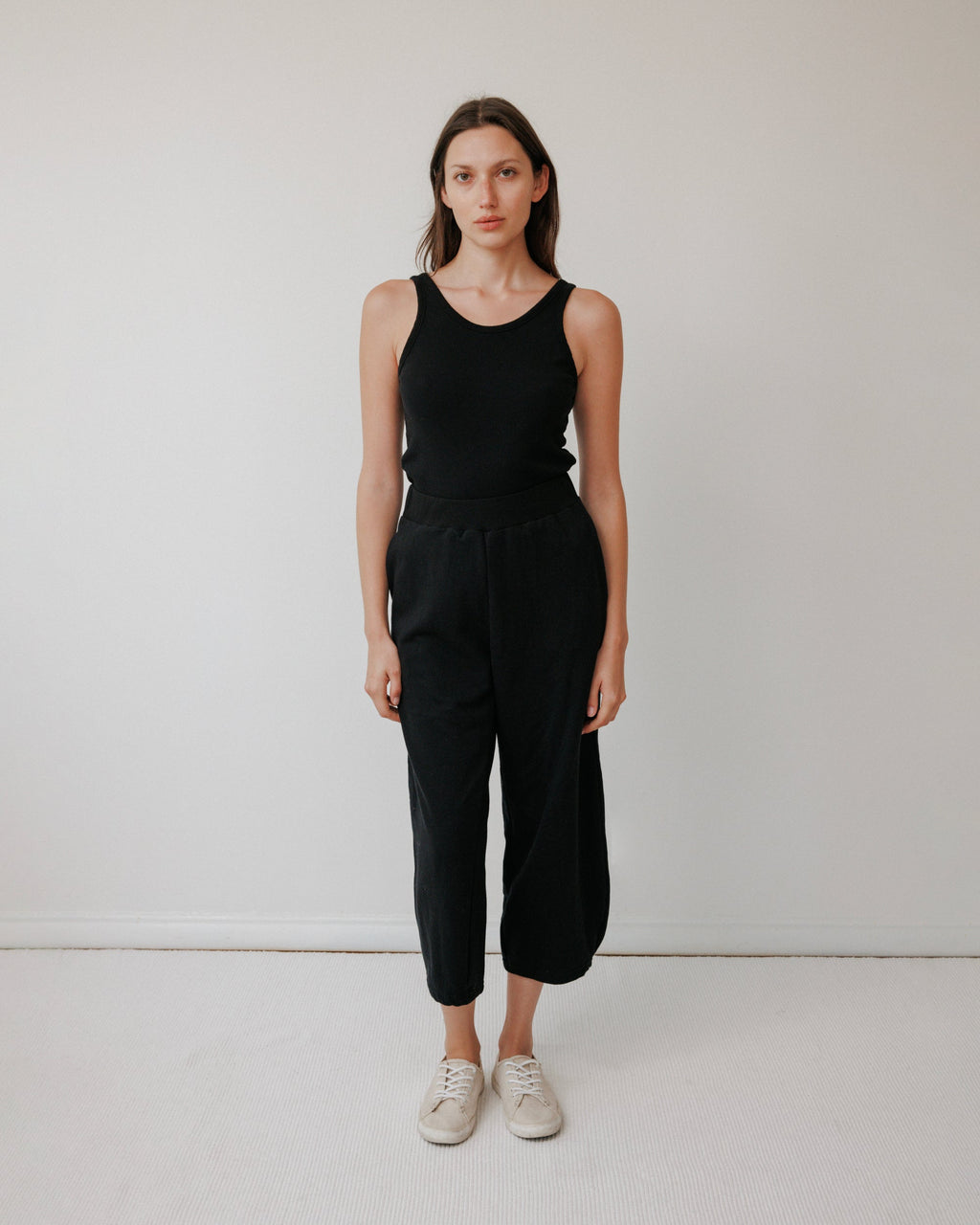 A woman stands against a white wall, wearing the Wol Hide Scoop Back Rib Tank in black, paired with black pants and white sneakers. She has straight brown hair, a neutral expression, and her arms are relaxed by her sides.