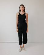 A woman stands against a white wall, wearing the Wol Hide Scoop Back Rib Tank in black, paired with black pants and white sneakers. She has straight brown hair, a neutral expression, and her arms are relaxed by her sides.