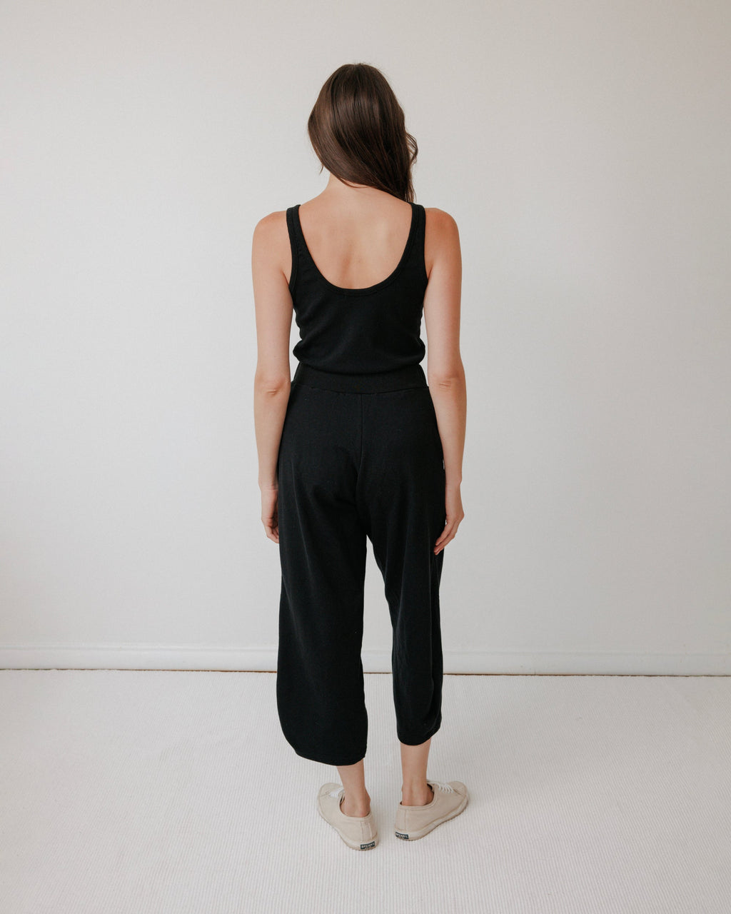 A woman with long brown hair wears the Wol Hide Scoop Back Rib Tank in black, paired with black pants and beige slip-on shoes, standing facing away from the camera against a plain white wall.