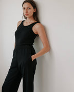 A woman with long brown hair models the Wol Hide Scoop Back Rib Tank in black, paired with black pants. She stands against a plain white wall, hands in her pockets, looking calmly at the camera.
