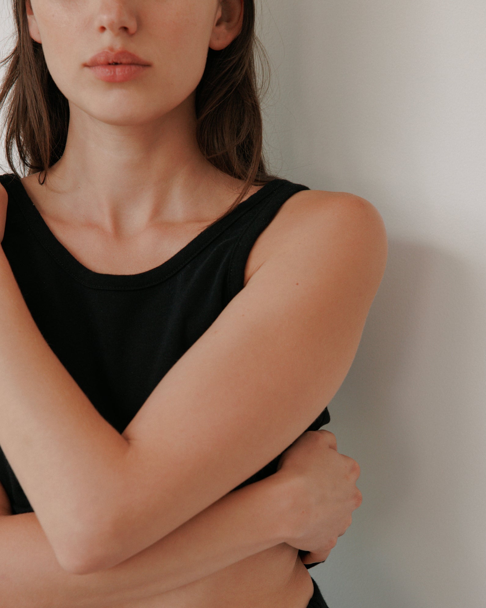 A woman with long brown hair wears the Wol Hide Scoop Back Rib Tank in black, standing against a white wall with arms crossed and a neutral, partially visible expression.
