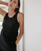 Wearing the Wol Hide Scoop Back Rib Tank in Black with matching pants, a person leans against a white wall, gazing at the camera with one hand behind their head. A window showing green trees is visible in the background.