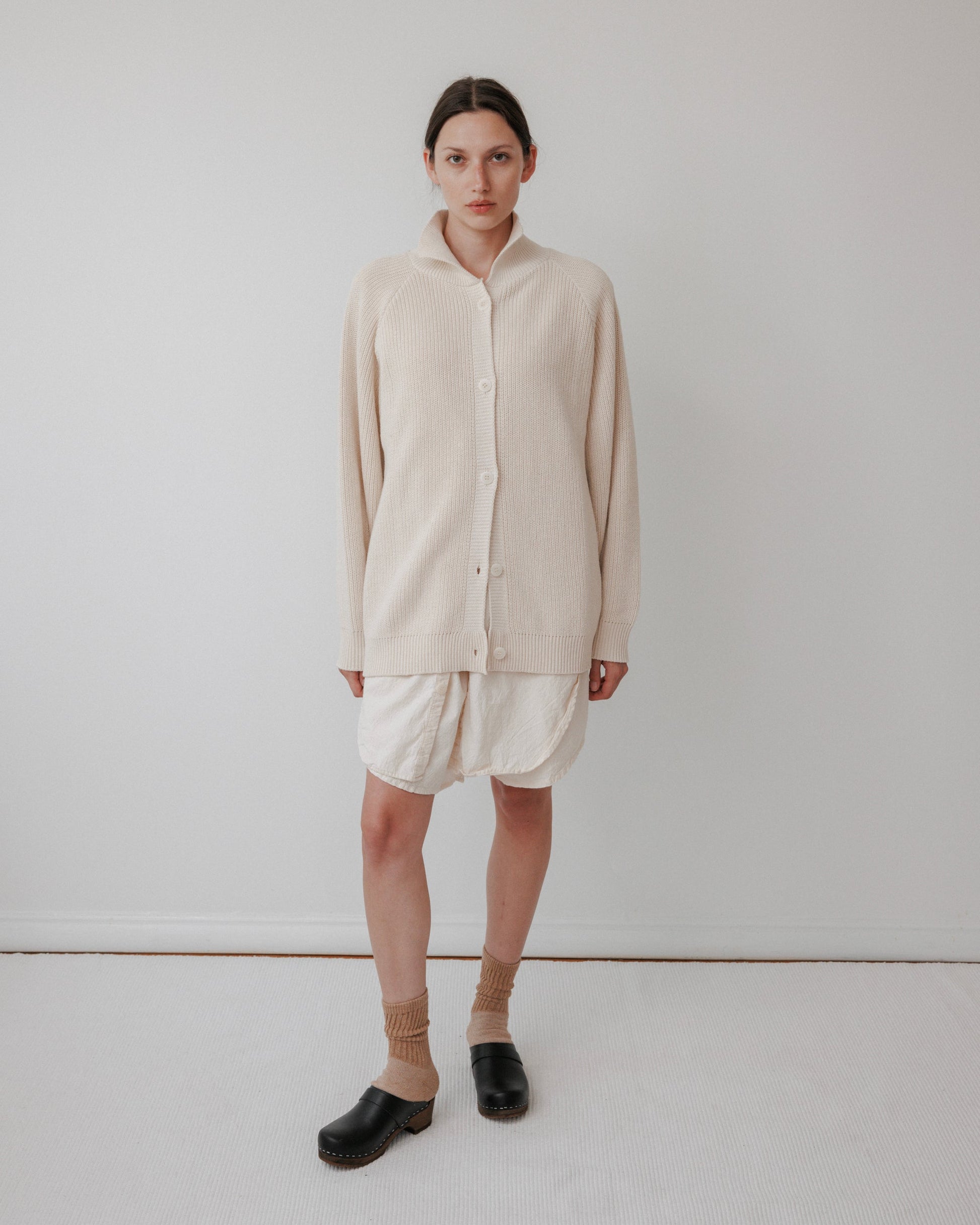 A person stands in a minimalist room wearing Wol Hide's Collar Cardigan: Natural, white shorts, tan socks, and black clogs. The cardigan's textures beautifully complement the plain white wall and floor, enhancing a clean and simple aesthetic.