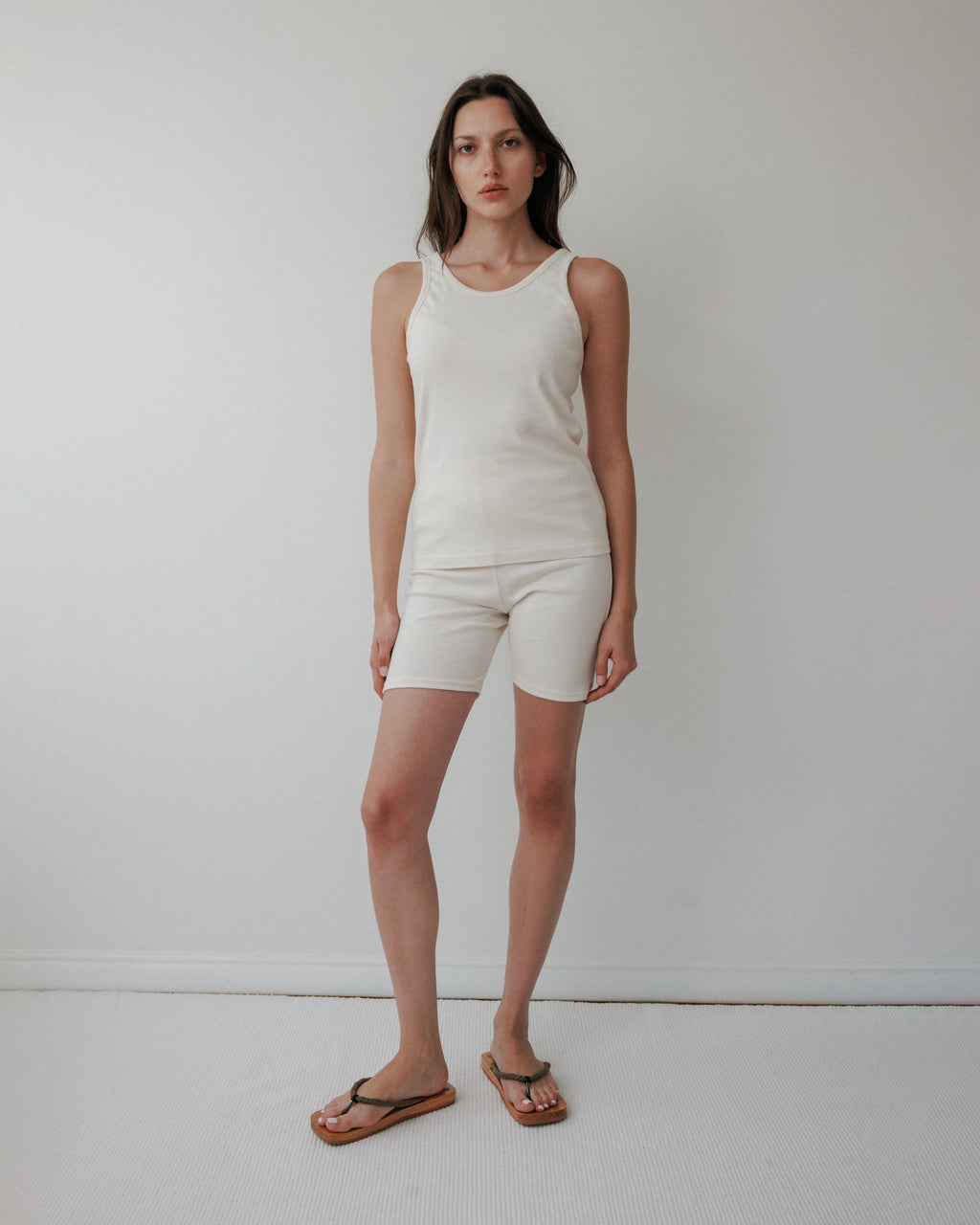 A woman with long brown hair and a neutral expression stands against a plain white wall, wearing Wol Hide’s Scoop Back Rib Tank in Natural and matching beige shorts, paired with brown flip-flops. The scene is minimal and well-lit.