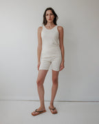A woman with long brown hair and a neutral expression stands against a plain white wall, wearing Wol Hide’s Scoop Back Rib Tank in Natural and matching beige shorts, paired with brown flip-flops. The scene is minimal and well-lit.