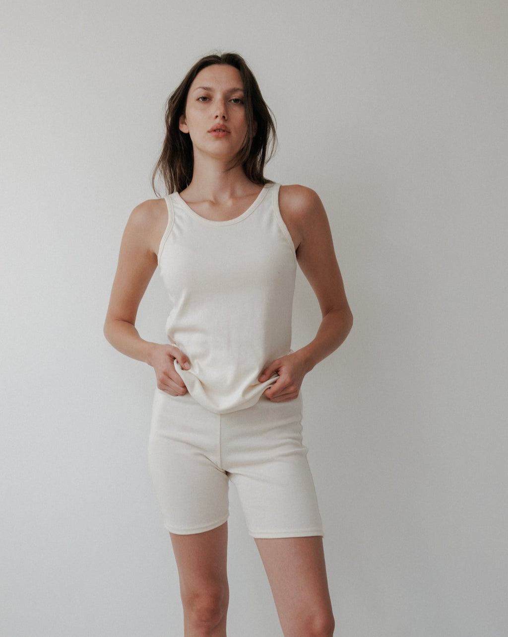 A woman stands against a white background wearing the Wol Hide Scoop Back Rib Tank in Natural, paired with matching shorts. She faces the camera with a neutral expression, hands resting on her top.