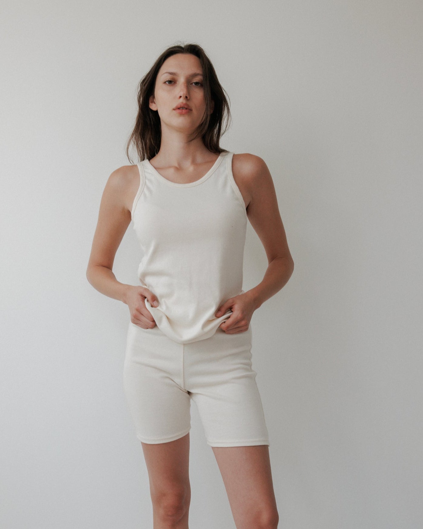 A woman stands against a white background wearing the Wol Hide Scoop Back Rib Tank in Natural, paired with matching shorts. She faces the camera with a neutral expression, hands resting on her top.