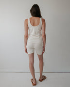 A woman with long brown hair stands facing a white wall, her back to the camera, wearing Wol Hide's Scoop Back Rib Tank in Natural paired with white shorts and brown sandals.