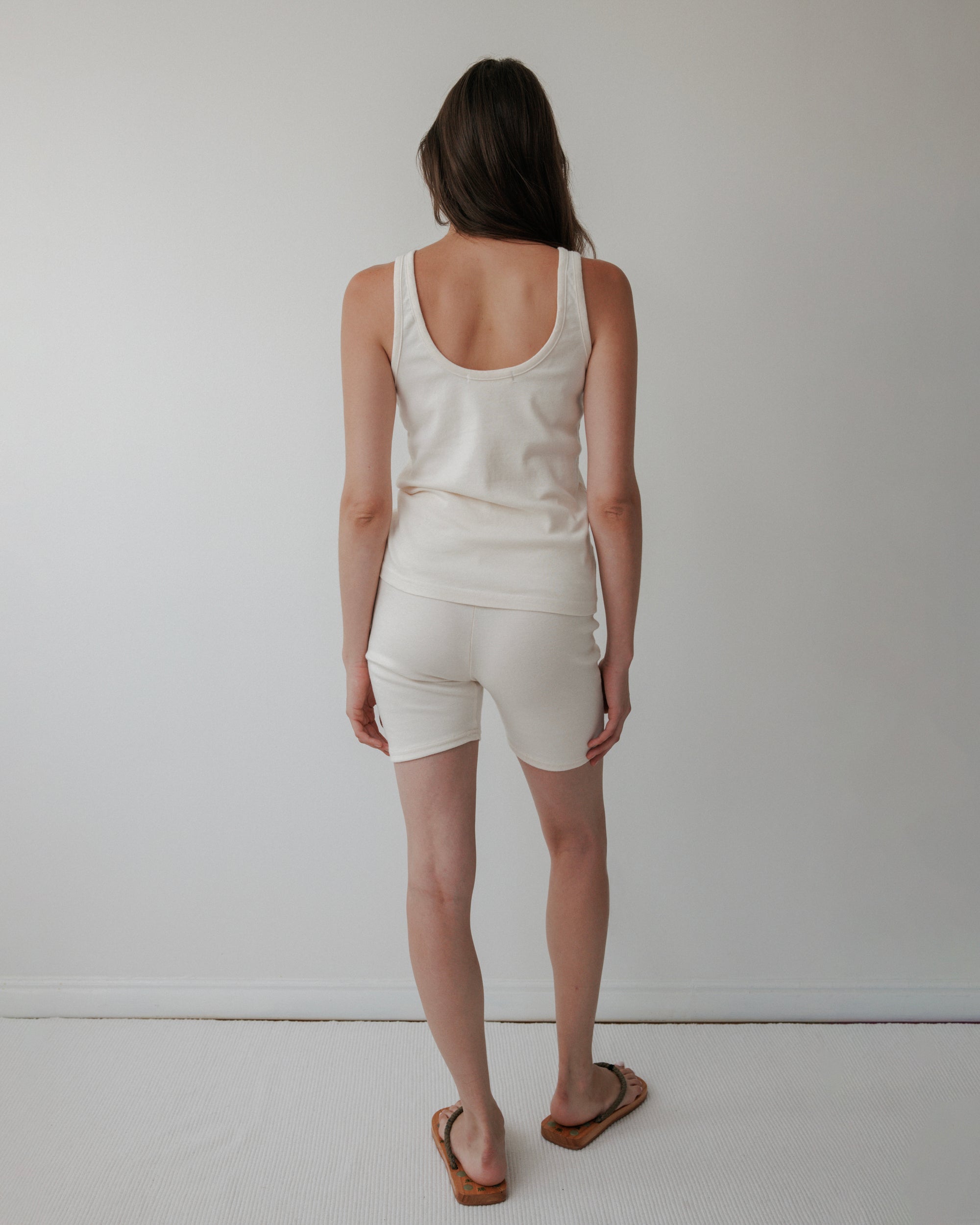 A woman with long brown hair stands facing a white wall, her back to the camera, wearing Wol Hide's Scoop Back Rib Tank in Natural paired with white shorts and brown sandals.