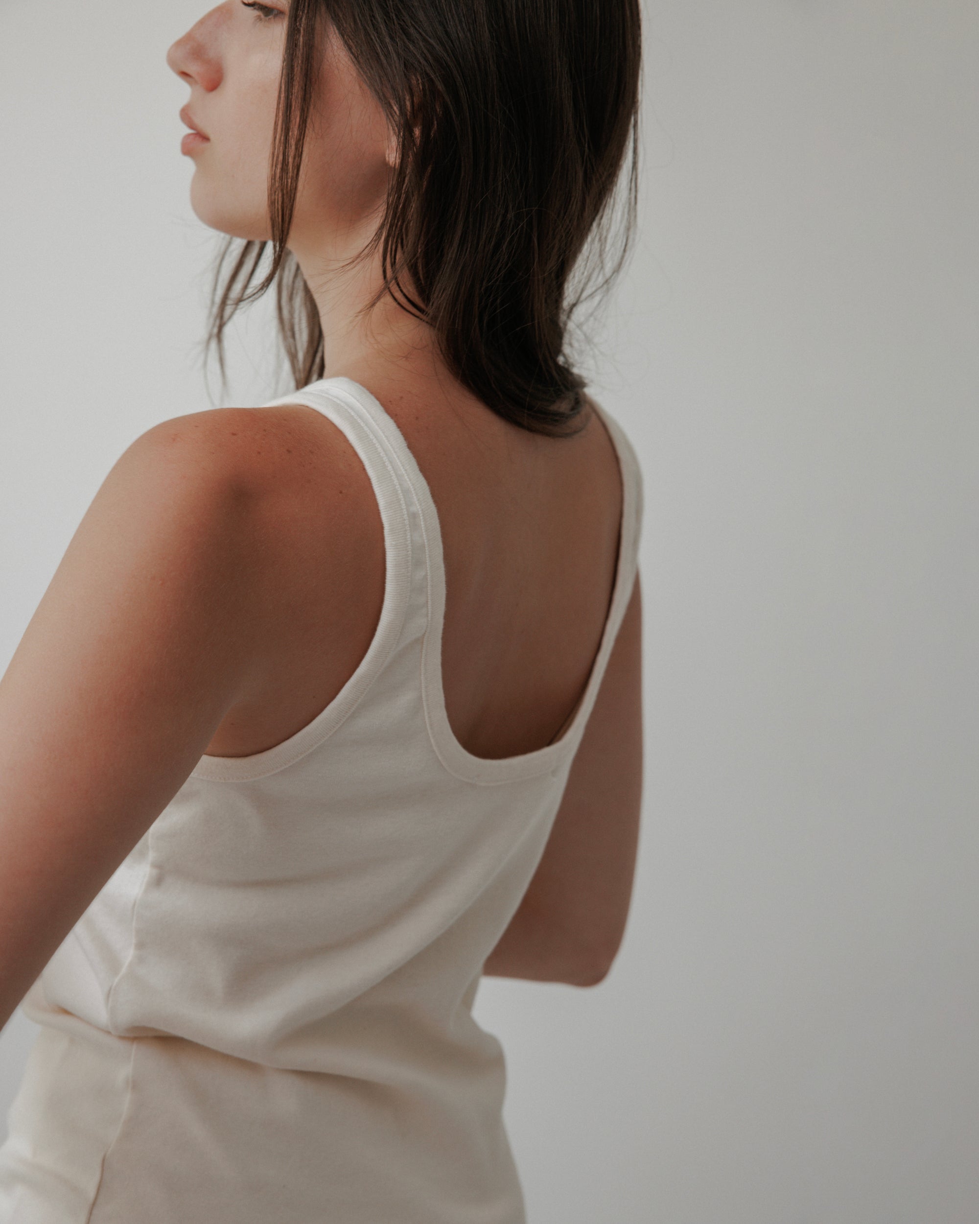 A woman with long brown hair models the Wol Hide Scoop Back Rib Tank in Natural, made from organic cotton, fitted to her frame and shown from the side and back against a plain light background. Her face is partially turned away.