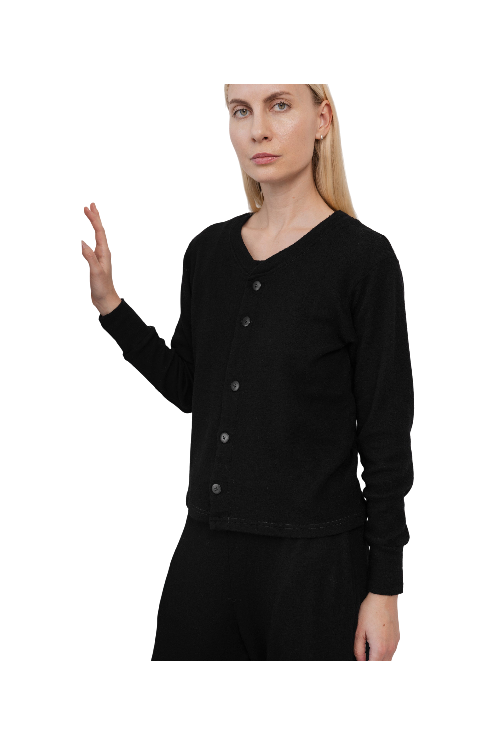 A woman with straight blonde hair models the Wol Hide Wool Liner in Onyx—a minimalist black cardigan—paired with black pants against a white background, showcasing a modern, sustainable style as she raises her left hand.