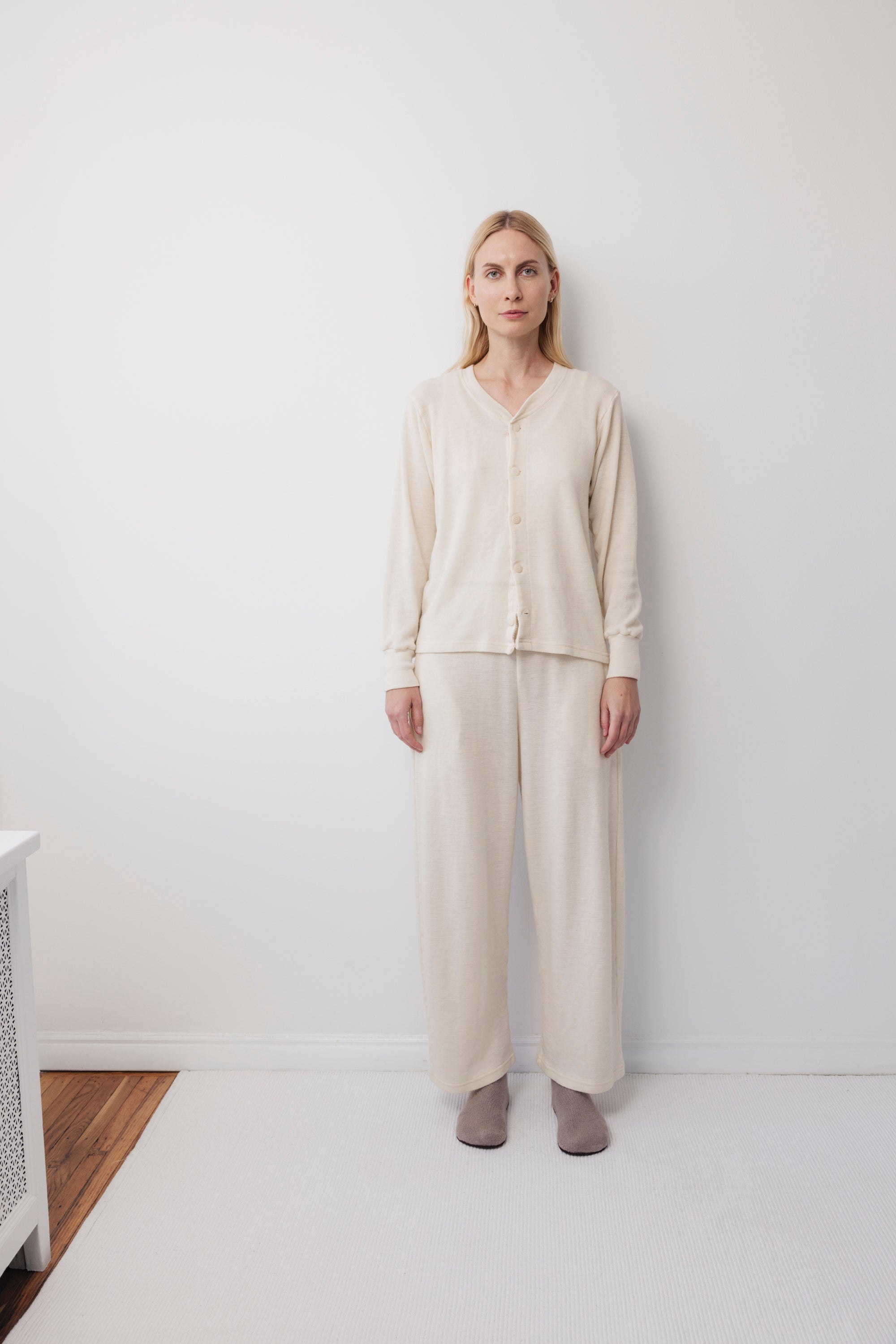 A woman stands against a plain white wall wearing Wol Hide's Wool Wide Leg: Natural pants with an elastic waist, a cream cardigan, and gray slippers. The floor and background remain minimal and uncluttered.
