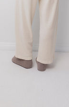 A person wearing Wol Hide's Wool Wide Leg: Natural pants and brown socks stands on a light carpet near a white wall, with only their lower legs and feet visible.