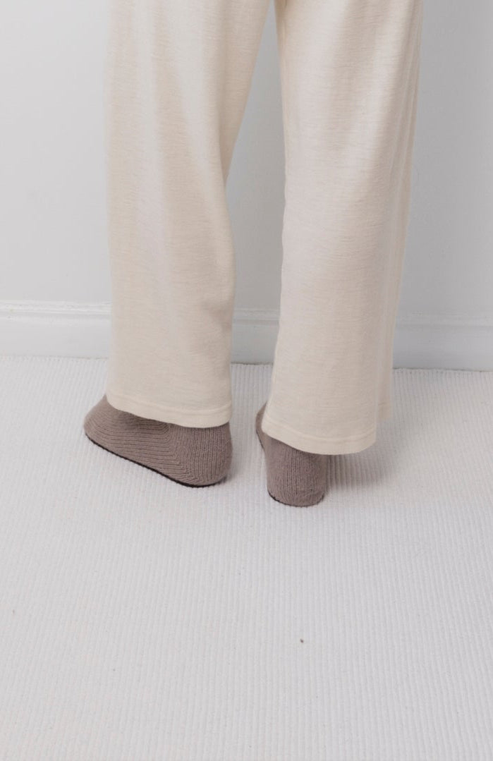 A person wearing Wol Hide's Wool Wide Leg: Natural pants and brown socks stands on a light carpet near a white wall, with only their lower legs and feet visible.