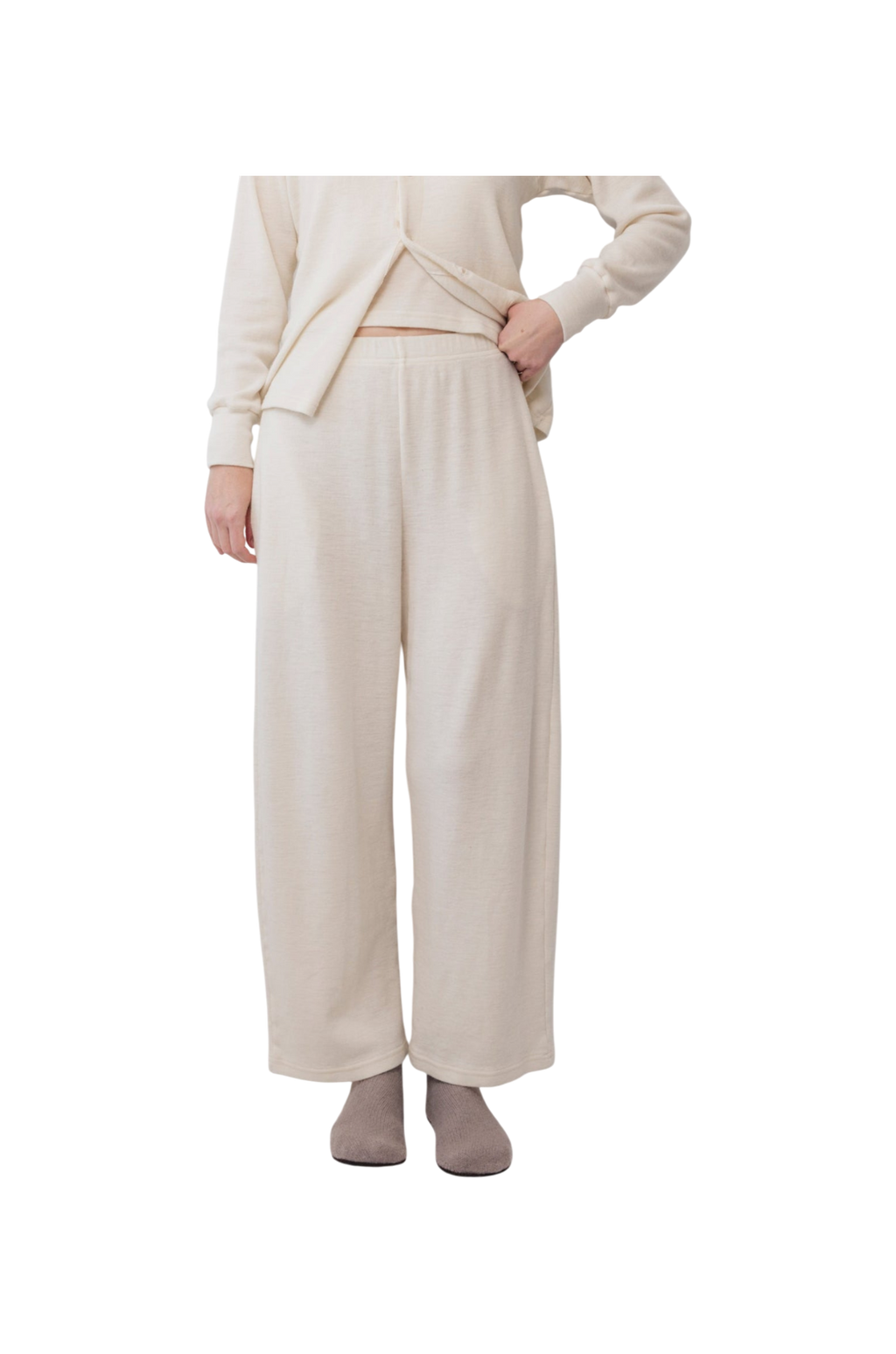 A person wears Wol Hide’s Wool Wide Leg: Natural lounge pants with a matching top and gray slippers, standing against a white background. Their hand is on their hip, and their face is out of frame.