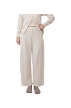 A person wears Wol Hide’s Wool Wide Leg: Natural lounge pants with a matching top and gray slippers, standing against a white background. Their hand is on their hip, and their face is out of frame.