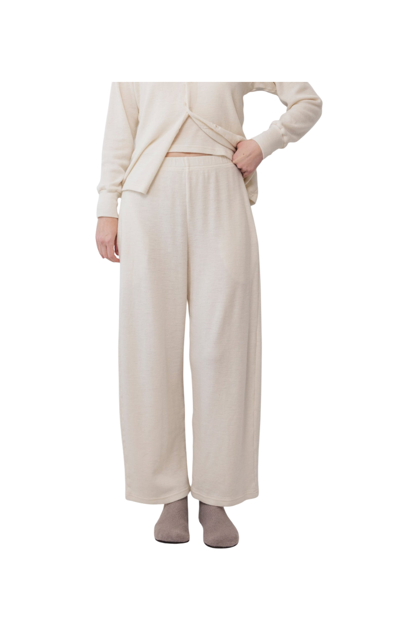 A person wears Wol Hide’s Wool Wide Leg: Natural lounge pants with a matching top and gray slippers, standing against a white background. Their hand is on their hip, and their face is out of frame.