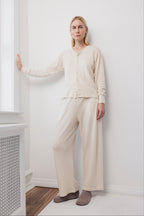 A woman stands indoors against a white wall, wearing the Wol Hide Wool Wide Leg pants in Natural, paired with a cream-colored cardigan and beige slippers. She has blonde hair and a calm expression, one hand resting on the wall beside a window.