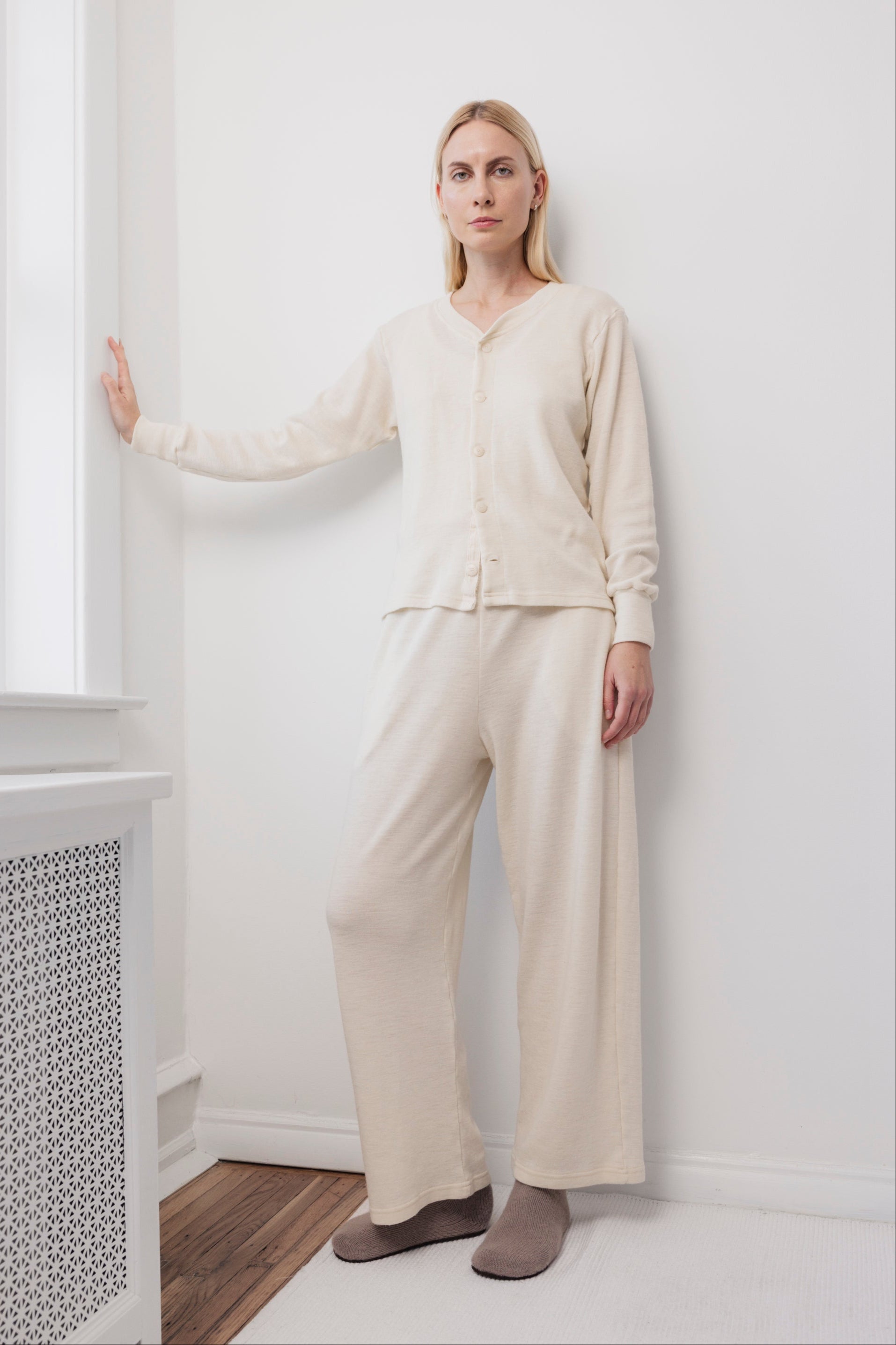 A woman stands indoors against a white wall, wearing the Wol Hide Wool Wide Leg pants in Natural, paired with a cream-colored cardigan and beige slippers. She has blonde hair and a calm expression, one hand resting on the wall beside a window.