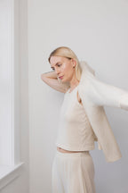 A blonde woman wears the Wol Hide Wool Tank in Natural with matching pants, adding a lightweight wool jacket while standing in a bright room with white walls and a window.