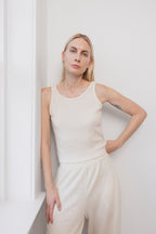 A woman with long blonde hair wears the Wol Hide Wool Tank in Natural, paired with matching pants. She stands by a white wall, one hand on the windowsill and the other on her hip, highlighting the chic scoop back detail.