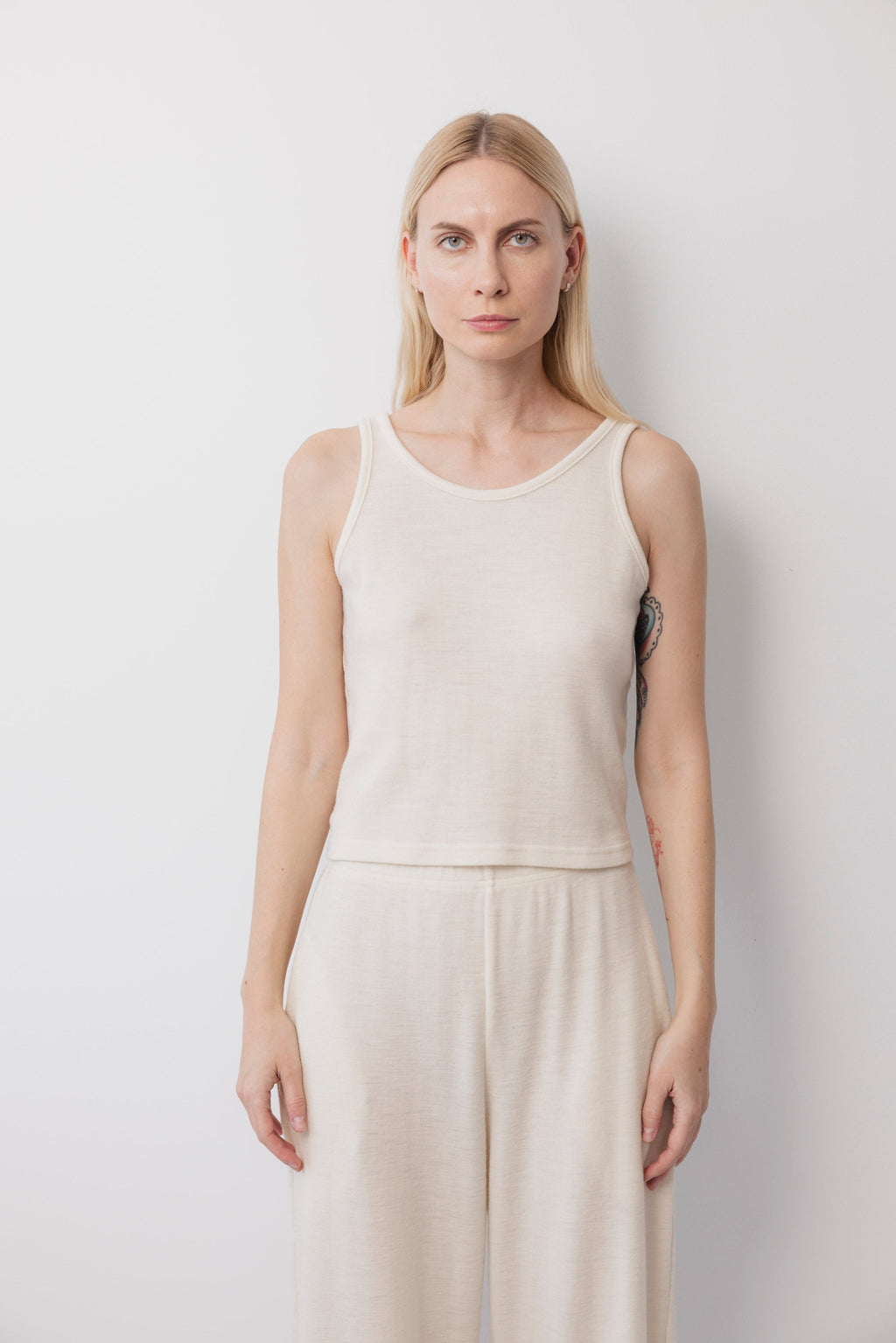 A woman with straight blonde hair wears the Wol Hide Wool Tank in Natural, paired with matching wide-leg pants. She stands against a plain white background facing forward, showing a colorful tattoo partially visible on her left side.