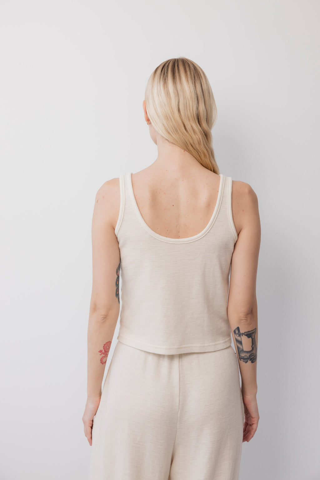 A person with long blonde hair and visible arm tattoos faces away from the camera, wearing Wol Hide’s Wool Tank: Natural in light beige wool. The setting is a plain white background.