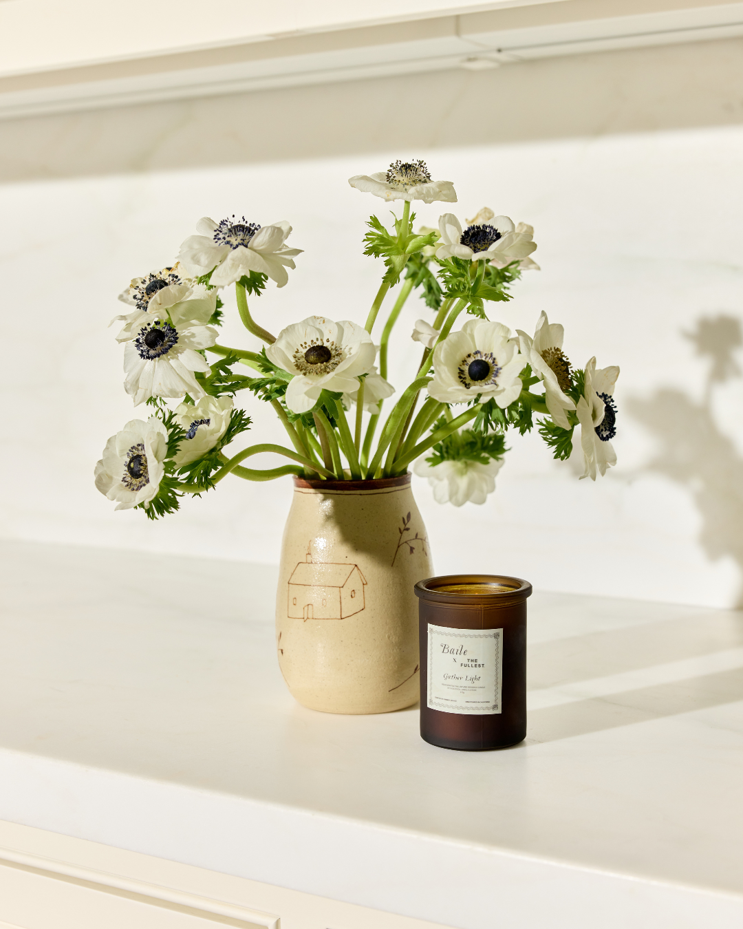 The Baile Homestead Vase by Baile, filled with white anemones, sits on a white countertop beside a brown "Field" candle jar. Sunlight casts soft shadows in the background.