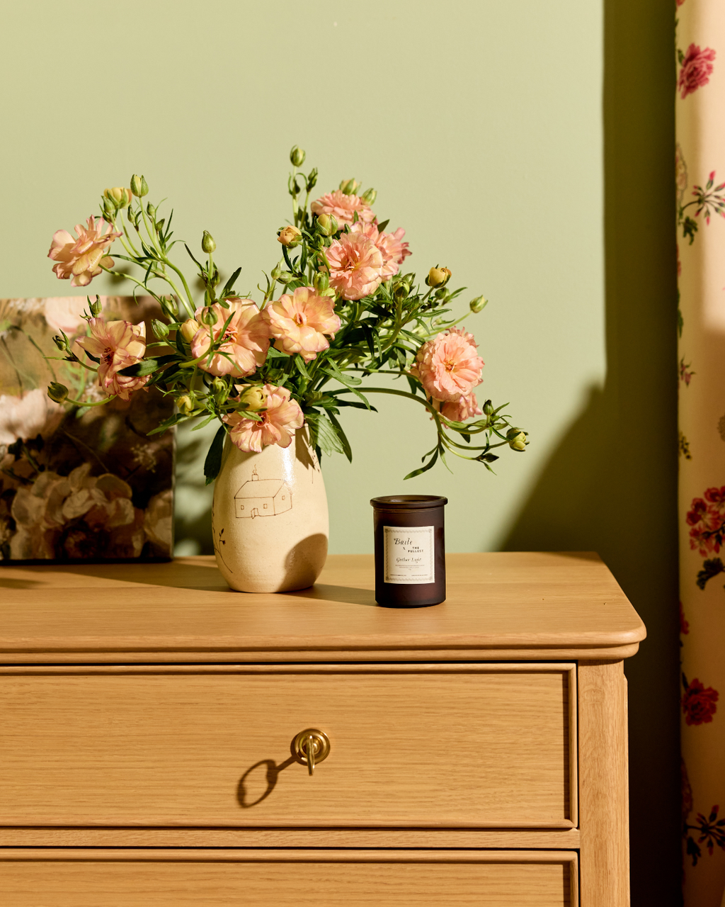 A wooden dresser with a key in the drawer features the Baile Homestead Vase by Baile, filled with pink flowers, and a dark candle on top. The setup sits against a green wall with floral curtains lit by gentle sunlight.