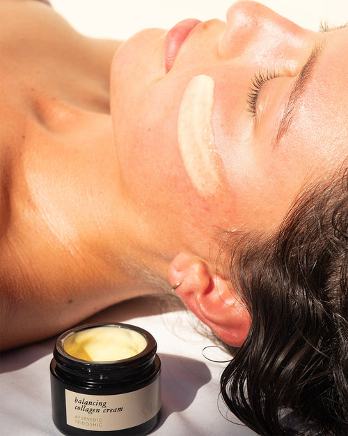 A woman relaxes in sunlight with her eyes closed, a creamy stripe of Surya Balancing Collagen Cream on her cheek. In the foreground, an open jar labeled "Balancing Collagen Cream" sits on a white surface.