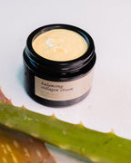 A black Surya Balancing Collagen Cream jar, labeled "Ayurvedic, Tridoshic," sits open to reveal its creamy texture. An aloe vera leaf lies diagonally in front, highlighting the product’s herbal support and skin-balancing benefits.