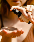A person dispenses Surya Balancing Face Oil from a dark glass bottle into their palm. They wear gold rings, have light brown hair, and the softly blurred background emphasizes the nourishing Ayurvedic ritual.