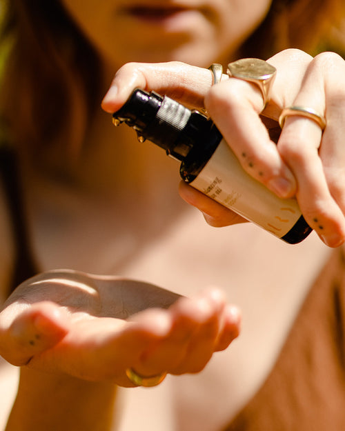 A person dispenses Surya Balancing Face Oil from a dark glass bottle into their palm. They wear gold rings, have light brown hair, and the softly blurred background emphasizes the nourishing Ayurvedic ritual.