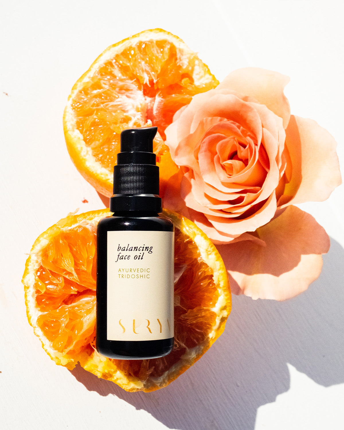 A bottle of Surya Balancing Face Oil sits atop a halved orange, accompanied by an orange rose, all displayed against a bright white background.