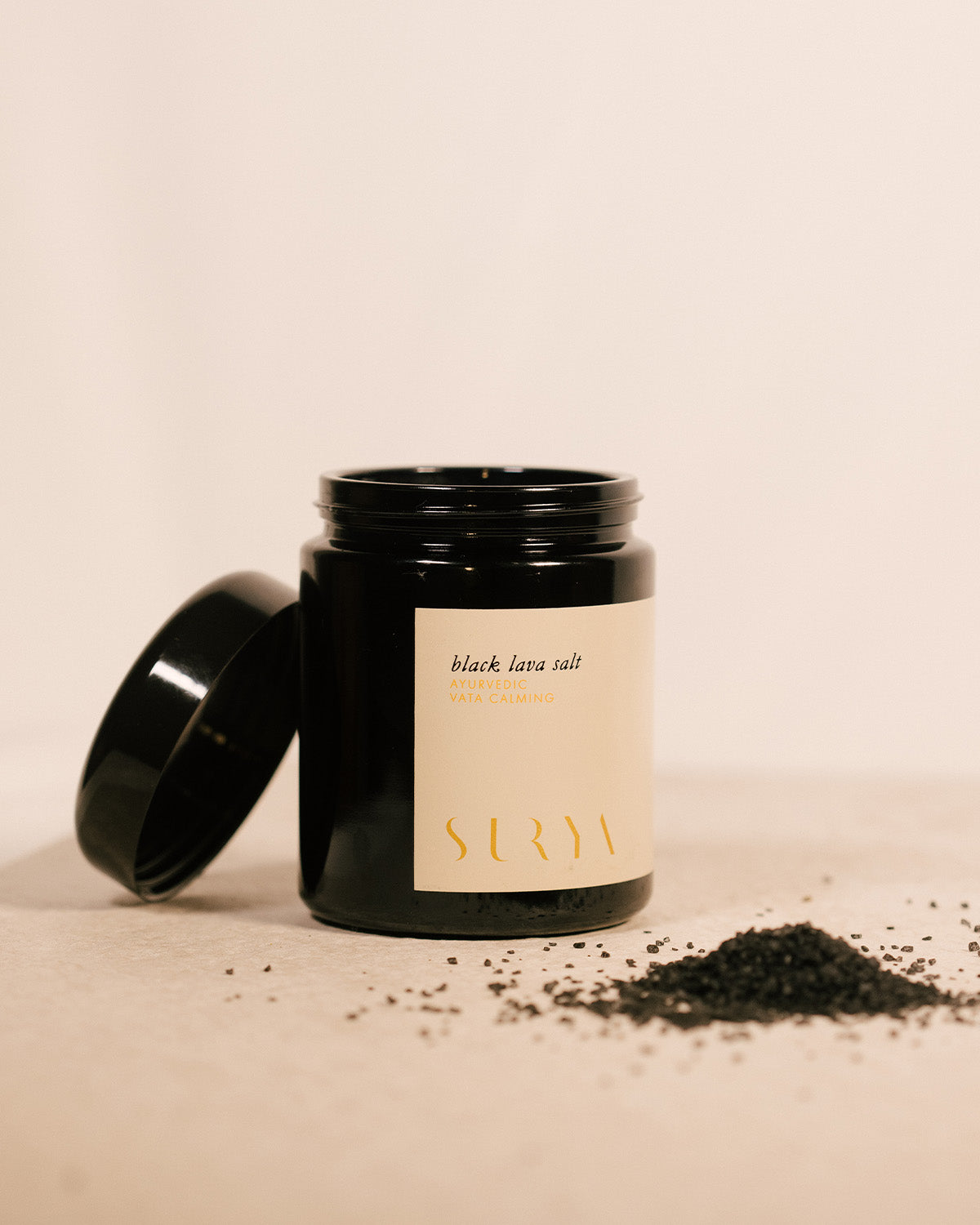 A black jar labeled "Black Lava Salt" by Surya sits open on a neutral surface, with Hawaiian salt blended with activated charcoal spilled beside it.
