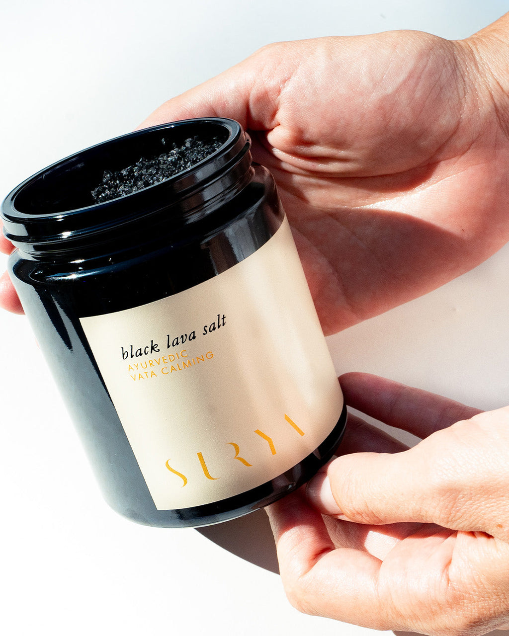 A hand holds an open black jar of Surya Black Lava Salt, showing coarse salt inside. The label reads "Black Lava Salt." Bright light casts shadows on the white background.
